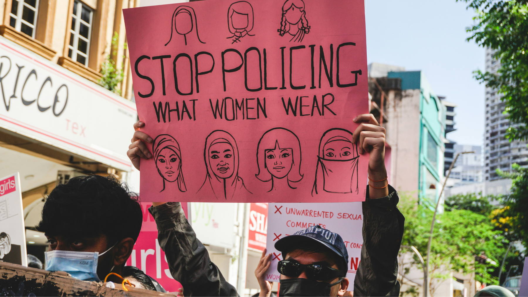 Image of two men protesting with a sign that says ““Stop policing what women wear” while avoiding facial recognition by wearing a face mask, sunglasses and cap.